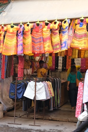A vibrant street market stall featuring an array of colorful dresses hanging prominently at the front. The dresses are primarily in shades of orange, yellow, red, and blue, with intricate patterns. Beneath the dresses, there are racks of shorts and more clothing items. Two people are visible inside the stall, adding a lively human element to the scene.