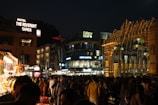 A bustling nighttime scene in an urban area, featuring a crowded street with people walking and shopping. Numerous brightly lit shops and a hotel with signage such as 'The Reverent Ganga' and 'Curry Leaf' are visible. Decorative string lights adorn the structures, enhancing the lively atmosphere.