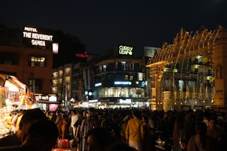 A bustling nighttime scene in an urban area, featuring a crowded street with people walking and shopping. Numerous brightly lit shops and a hotel with signage such as 'The Reverent Ganga' and 'Curry Leaf' are visible. Decorative string lights adorn the structures, enhancing the lively atmosphere.