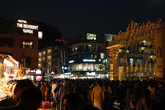 A bustling nighttime scene in an urban area, featuring a crowded street with people walking and shopping. Numerous brightly lit shops and a hotel with signage such as 'The Reverent Ganga' and 'Curry Leaf' are visible. Decorative string lights adorn the structures, enhancing the lively atmosphere.