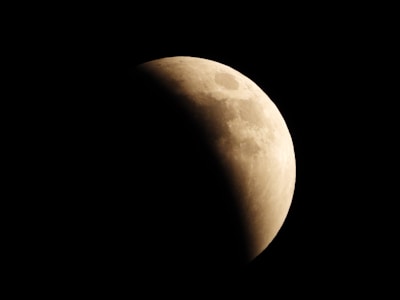 A partially shadowed moon with its illuminated portion in contrast against a dark sky. The surface of the moon displays visible craters and texture, showcasing details of lunar geography.