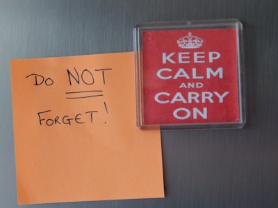 A sticky note with the hand-written message 'Do NOT Forget!' is attached to a vertical surface next to a magnet with the iconic phrase 'KEEP CALM AND CARRY ON' in white letters on a red background.