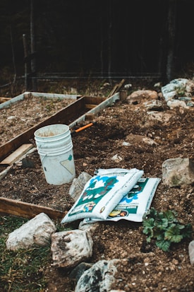 A garden area with raised beds containing soil. There are scattered rocks around, a white bucket with a maple leaf design, and a couple of bags of sheep manure lying on the ground. The area appears to be prepared for planting, with some small green plants visible.