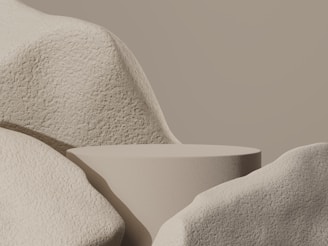 A minimalist composition with a smooth, rounded platform surrounded by large, textured stone-like forms. The monochromatic palette emphasizes the soft curves and shadows of the objects, creating a serene and balanced appearance.