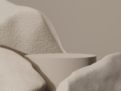 A minimalist composition with a smooth, rounded platform surrounded by large, textured stone-like forms. The monochromatic palette emphasizes the soft curves and shadows of the objects, creating a serene and balanced appearance.