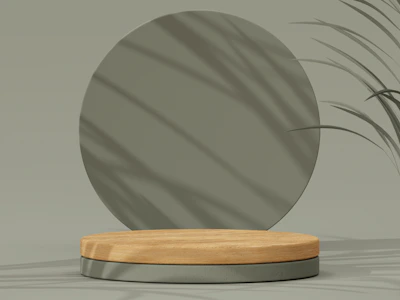 A minimalist composition featuring a round wooden platform set against a muted green backdrop. Subtle shadows from leaves fall across the surface, adding depth and texture to the scene.