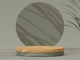 A minimalist composition featuring a round wooden platform set against a muted green backdrop. Subtle shadows from leaves fall across the surface, adding depth and texture to the scene.