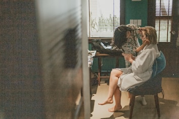 A woman sitting on a chair is having her makeup done by another person. The room has a relaxed atmosphere, with a window in the background allowing natural light in. The woman is wearing a white robe, and there are various cosmetic products on the nearby table.