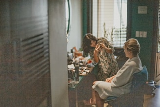 A woman in a white robe sits in a chair, likely getting her makeup done. Another person stands nearby with a phone, possibly taking a photo or checking something. The setting appears to be a room with dim lighting and assorted makeup items visible on a table.