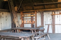 A rustic, wooden interior with benches and tables. A wooden ladder leans against a wall adorned with pottery on shelves. The setting includes woven baskets and other rustic decor, creating a historical or traditional atmosphere.