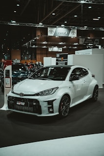A white sports car is prominently displayed in a showroom or car exhibition setting. The car has a sleek and modern design with aerodynamic features and black alloy wheels. The background includes banners and signs for various car brands like Toyota and Jaguar Land Rover, indicating a commercial or promotional event.