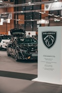 A car exhibition showing a black Peugeot vehicle with the hood open. Several people are visible in the background, and signage from car brands Peugeot and Renault can be seen suspended above the exhibition area. A prominent Peugeot logo and information board are located in the foreground.