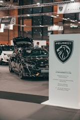 A car exhibition showing a black Peugeot vehicle with the hood open. Several people are visible in the background, and signage from car brands Peugeot and Renault can be seen suspended above the exhibition area. A prominent Peugeot logo and information board are located in the foreground.