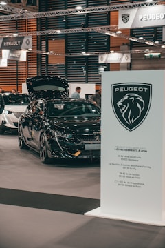 A car exhibition showing a black Peugeot vehicle with the hood open. Several people are visible in the background, and signage from car brands Peugeot and Renault can be seen suspended above the exhibition area. A prominent Peugeot logo and information board are located in the foreground.