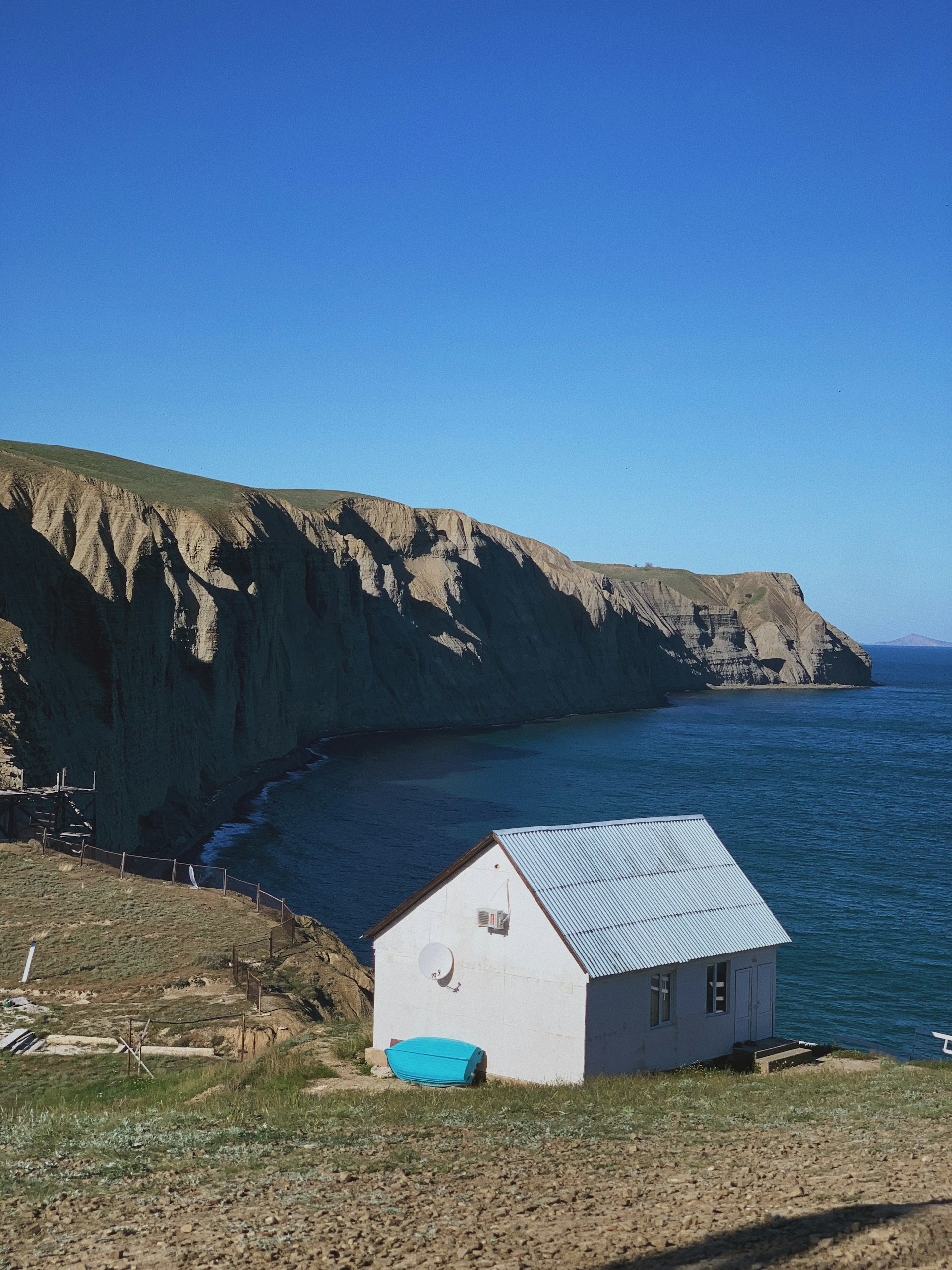A small white house with a satellite dish and blue roof sits near the edge of a cliff overlooking a vast blue sea. The coastline is rugged, with steep cliffs and green-topped hills. The sky is clear and blue, enhancing the serene and remote setting.
