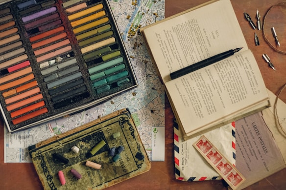 A wooden surface is covered with various artistic and vintage items. A box of colorful pastel chalks is prominently displayed next to an open book with text. Vintage postage stamps and envelopes are scattered around, along with several metal pen nibs. A map is partially visible underneath the other objects.