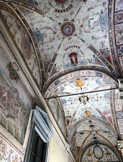 Intricate frescoes adorn a vaulted ceiling, featuring detailed historical and mythical scenes with an array of figures and decorative motifs. The adjacent wall showcases a large mural painting likely depicting a historical cityscape. The rich detail and artistic style suggest a Renaissance influence, characterized by ornate patterns and vibrant imagery.