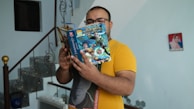 A person wearing glasses and a yellow shirt is reading a colorful comic book with superhero characters on the cover. The setting appears to be indoors with a staircase in the background and a framed portrait on the wall.