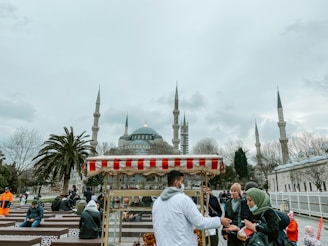 A scene in front of a large mosque with multiple minarets and a central dome. Several people are gathered near a simit cart. The weather appears overcast with a cloudy sky, and there are trees without leaves in the background.