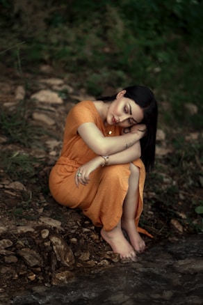 A woman wearing an orange dress sits on rocky ground near a stream, resting her head on her arm with closed eyes. The setting is lush with greenery, suggesting a tranquil, natural environment.