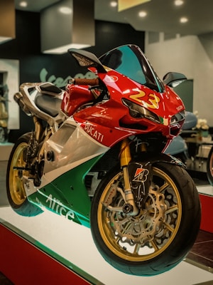 A sleek racing motorcycle with a red, white, and green color scheme. The bike has prominent branding with the number 23 on the front and the name 'Ducati' visible. It features golden wheel rims and is displayed on a platform in a showroom with soft lighting.