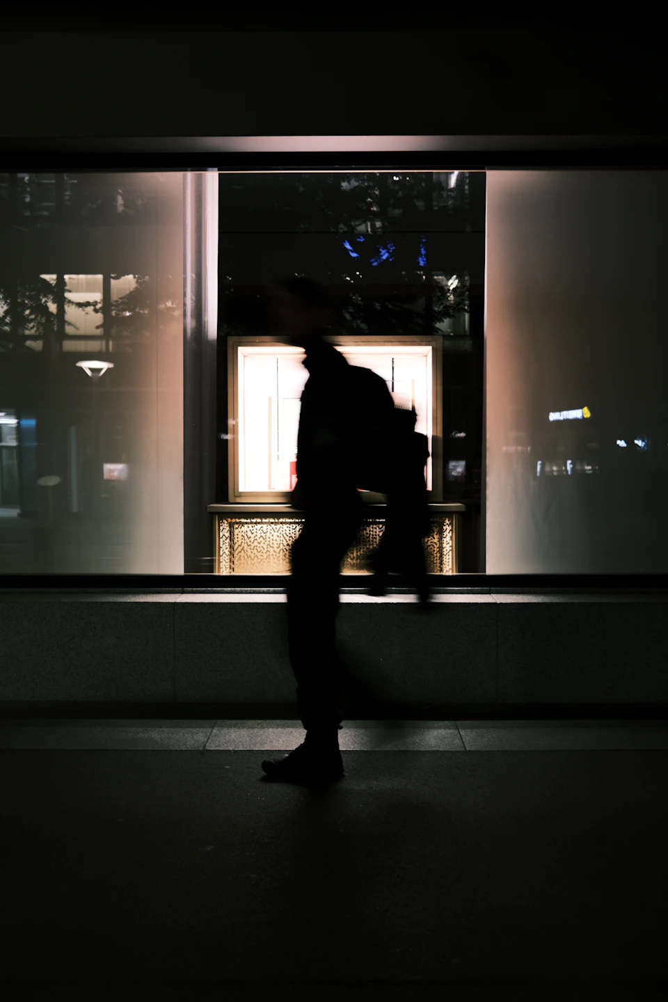 A lone figure walks on a sidewalk at night, silhouetted against store window lights