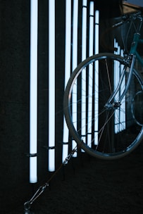 A bicycle is partially visible with a large focus on its front wheel, which is positioned against a background of vertical white LED light tubes. The light creates a stark contrast against the dark surroundings, highlighting the metal and rubber of the bicycle components.