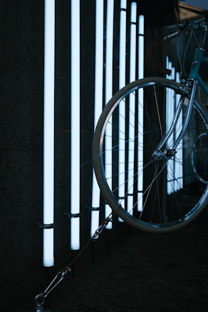 A bicycle is partially visible with a large focus on its front wheel, which is positioned against a background of vertical white LED light tubes. The light creates a stark contrast against the dark surroundings, highlighting the metal and rubber of the bicycle components.