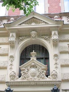 An elaborate architectural facade features ornate carvings, including three sculpted faces and intricate detailing around an arched window. Above the window, there's a crest with the year 1886 inscribed. Netting is visible to protect the structure, possibly to prevent birds from nesting. The facade includes decorative elements reminiscent of classical architecture.