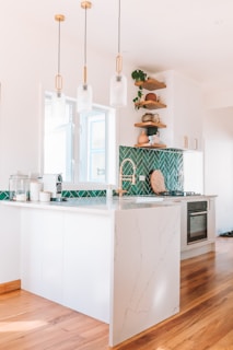 A modern kitchen with white cabinetry and a marble countertop. The green herringbone tile backsplash adds a vibrant touch. There are wooden shelves with decorative items and plants. Three pendant lights hang above the counter. The floor is wooden, and the kitchen appears bright and airy.