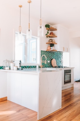 A modern kitchen with white cabinetry and a marble countertop. The green herringbone tile backsplash adds a vibrant touch. There are wooden shelves with decorative items and plants. Three pendant lights hang above the counter. The floor is wooden, and the kitchen appears bright and airy.