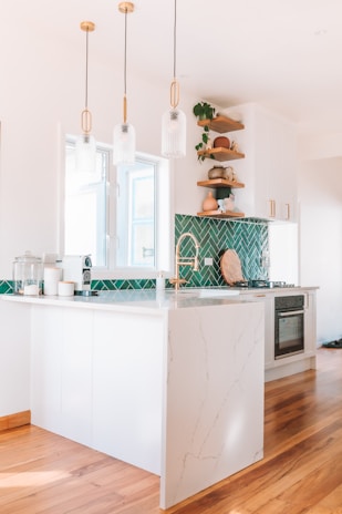 A modern kitchen with white cabinetry and a marble countertop. The green herringbone tile backsplash adds a vibrant touch. There are wooden shelves with decorative items and plants. Three pendant lights hang above the counter. The floor is wooden, and the kitchen appears bright and airy.