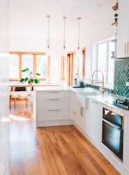 A modern kitchen with white cabinetry, wooden floors, and brass accents. There are three pendant lights hanging over the island. A large window allows natural light to flood the space. A plant is placed near the window, adding a touch of greenery.