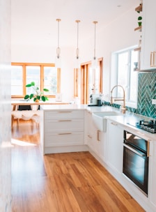 A modern kitchen with white cabinetry, wooden floors, and brass accents. There are three pendant lights hanging over the island. A large window allows natural light to flood the space. A plant is placed near the window, adding a touch of greenery.
