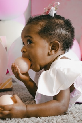 A young child lying on a soft carpet with balloons in the background. The child is wearing light-colored clothing with a decorative bow in their hair, holding a pink ball near their mouth.