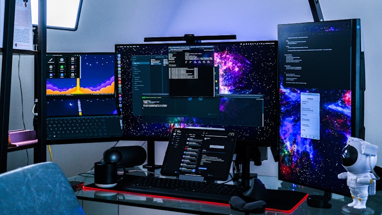 A multi-monitor computer setup with three screens displaying various software applications and cosmic-themed wallpapers. On the desk, there is a keyboard, a mouse, a tablet with a stand, a small speaker, and a white astronaut figurine. A chair is partially visible in the foreground.