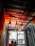 Hanging lights crafted from repurposed wine bottles illuminate a room with an industrial aesthetic. The ceiling features metal beams and distressed wood slats, casting a warm glow with orange and yellow hues. Below, two individuals face each other by a window that offers a view of an urban street.