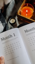 An open planner displays a calendar layout for 'Month 1' with weekly dates listed. In the background, there is a cozy setup with a lit orange candle in a wooden box and a smaller unlit white candle. A plaid blanket is partially visible, hinting at a warm, comfortable atmosphere.