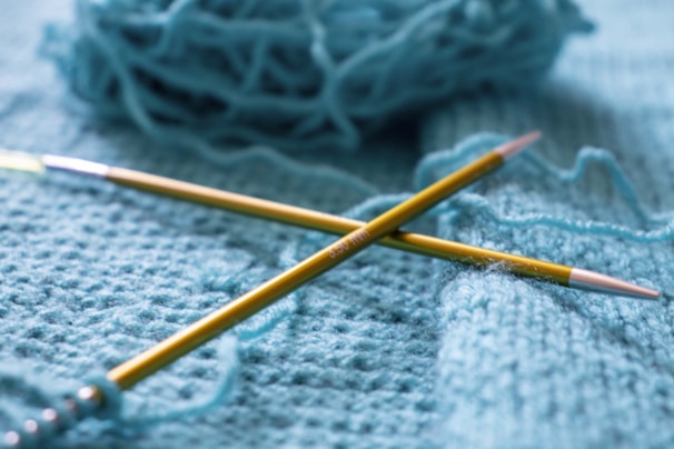 Two yellow knitting needles are crossed over a piece of knitted fabric. In the background, a ball of blue yarn is partially visible, creating a soft and cozy atmosphere.