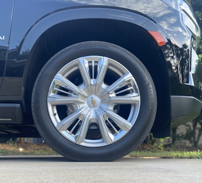 A close-up view of a car's shiny, silver alloy wheel with a Bridgestone tire. The background shows a blurred hint of greenery and the pavement.