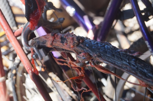 A close-up view of a severely damaged and melted bicycle tire attached to a frame. The rubber appears to be charred and deformed, with remnants hanging loosely. The frame is partially visible, displaying a red color, and there are hints of other bicycles in the background with purple components.