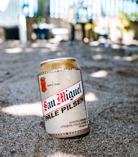 A can of San Miguel Pale Pilsen is placed on sandy ground with a blurred background of what appears to be a garden or outdoor area. The can has a classic design with red and black text against a gold and beige background.