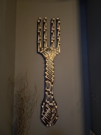 A large art piece composed of numerous metal forks meticulously arranged to form the shape of an oversized fork. The sculpture is mounted on a neutral-colored wall. To the left, dried branches add a natural element to the otherwise metallic and industrial composition.