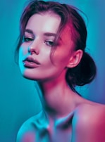 A portrait of a woman with her hair tied back and loose strands framing her face. The lighting creates a high-contrast effect with vibrant blue and pink hues, highlighting her smooth skin and glossy lips. Her facial expression appears calm and contemplative.