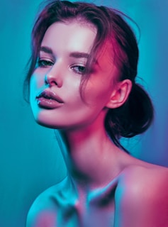 A portrait of a woman with her hair tied back and loose strands framing her face. The lighting creates a high-contrast effect with vibrant blue and pink hues, highlighting her smooth skin and glossy lips. Her facial expression appears calm and contemplative.