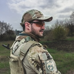 A person in camouflage military attire is outdoors in a field. The individual is wearing a tactical vest and a cap with a camo pattern, along with protective orange-tinted glasses. The background features overcast skies and sparse trees, suggesting a rural or wooded area.