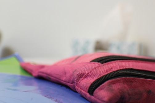A close-up view of a pink fabric pouch with black zippers, resting on a surface with some sheets or papers visible in the background.