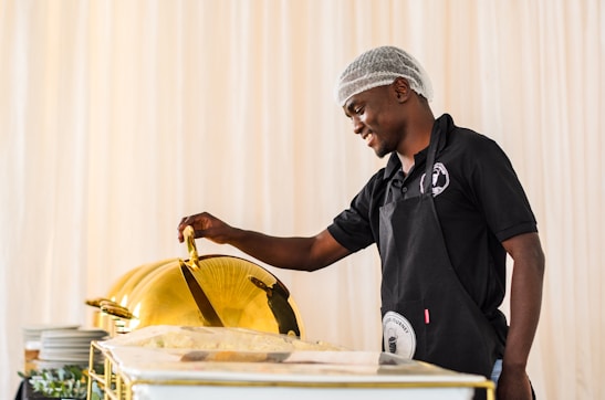 A person wearing a hairnet and dark polo shirt stands at a catering setup, lifting the lid of a chafing dish. Plates are stacked in the background, and the setting appears to be a professional or formal food service environment.