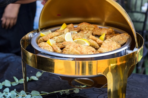 A golden chafing dish filled with crispy fried pieces of fish or chicken adorned with slices of fresh lemon. The setting suggests an elegant dining event, with the dish placed on a table covered in dark fabric.