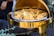 A golden chafing dish filled with crispy fried pieces of fish or chicken adorned with slices of fresh lemon. The setting suggests an elegant dining event, with the dish placed on a table covered in dark fabric.